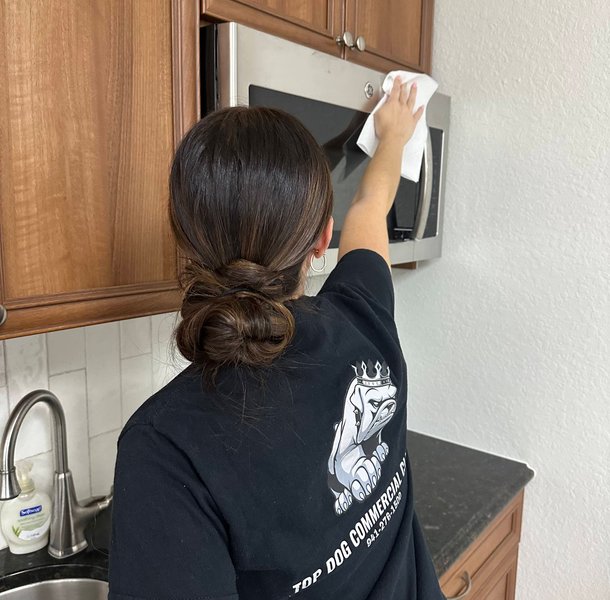 Woman wiping microwave hood in kitchen with wood cabinets.